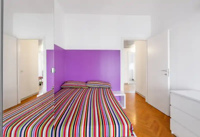 Camera da letto moderna e luminosa con letto a righe colorate e testata viola intensa. L'armadio specchiato amplifica lo spazio e il parquet dona calore.