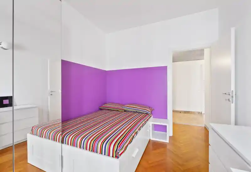 Camera da letto luminosa e moderna con letto bianco contro una parete viola, biancheria a righe e contenitori bianchi puliti — ben illuminata e arredata con cura per l'annuncio.