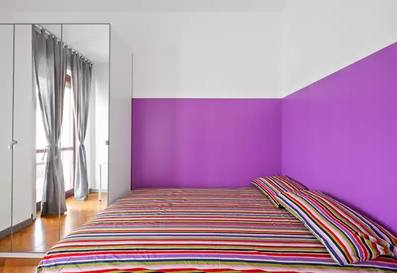 Camera da letto luminosa e moderna con un letto matrimoniale a righe colorate contro una parete viola vivace e un armadio con ante specchiate che si affaccia su un balcone — pulita, ben illuminata e di stile.