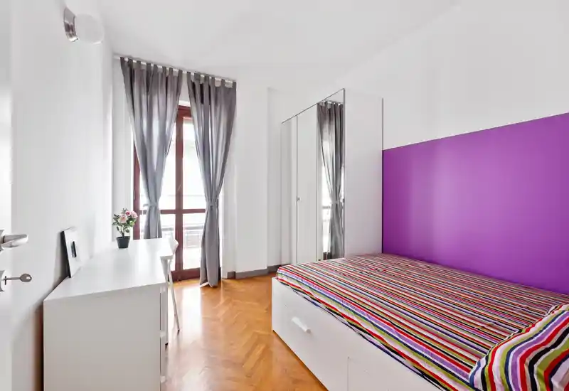 Camera da letto luminosa e moderna con letto a righe colorate, parete accentuata viola e armadio con ante a specchio. La luce naturale entra dalla porta del balcone con tende leggere.