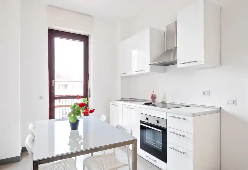 Bright modern kitchen with integrated oven, sleek white cabinets and a glass dining table by a tall window; simple, airy and well-lit space ideal for rental photos.