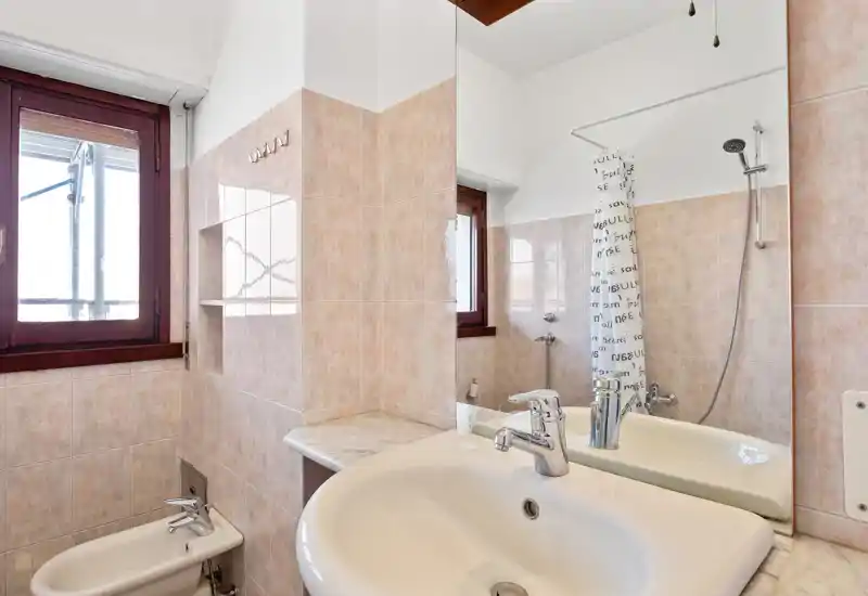 Clean, well-lit bathroom with a white sink, bidet and shower visible in the mirror; neutral beige tiles and a wooden-framed window provide natural light.