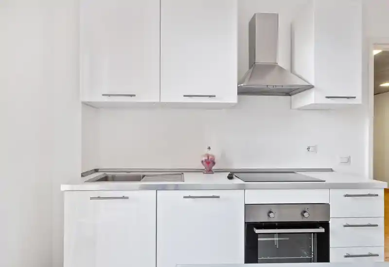 Modern compact kitchen with white cabinets, stainless-steel oven and extractor hood, integrated sink and induction hob. Bright, minimal and well-maintained space suitable for small apartments.