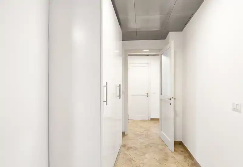 Bright, narrow entrance corridor with built-in white wardrobes and a light stone floor, leading to interior doors. Clean minimalist finishes and good natural/ambient lighting make this a functional entryway.