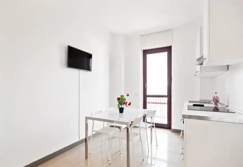 Bright, minimal kitchen with a small dining table and four chairs, wall-mounted TV and a balcony/door letting in natural light — neat and modern studio-style kitchen.