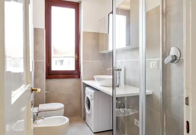Bright, modern bathroom with shower enclosure, toilet, bidet, washbasin and a compact washing machine neatly installed under the countertop.