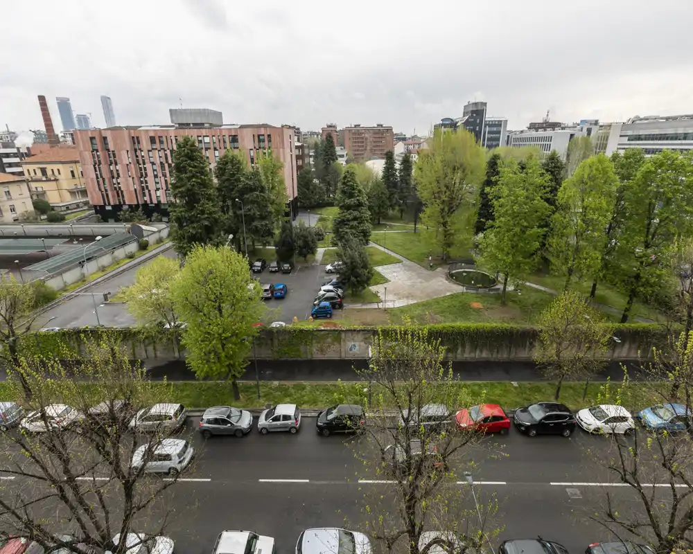 Vista desde altura de una calle con coches aparcados y un parque arbolado con caminos; cielo nublado y entorno urbano visible.