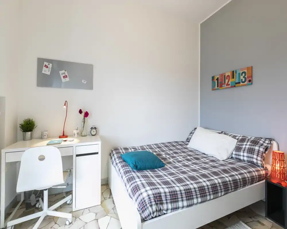 Dormitorio luminoso y moderno con cama doble con ropa de cuadros, cojín turquesa y una pequeña lámpara de noche. Un escritorio blanco compacto con silla y detalles decorativos hace la habitación práctica para trabajar y relajarse.
