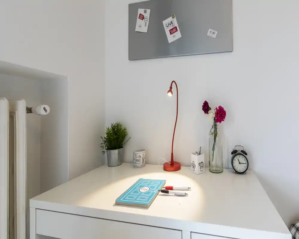Rincón luminoso y minimalista junto a la cama/escritorio con una pequeña lámpara de lectura roja, flores en jarrón, reloj despertador y cuaderno; un espacio ordenado y bien iluminado para leer o trabajar.