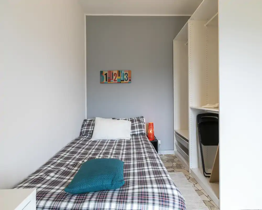 Dormitorio compacto y bien iluminado con cama individual con ropa de cuadros, una manta turquesa y una lámpara de mesita; armario abierto a la derecha con espacio de almacenamiento visible.