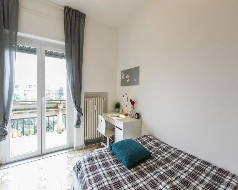Dormitorio luminoso y ordenado con puerta al balcón, un pequeño escritorio blanco y una cama con edredón de cuadros y cojín verde azulado.