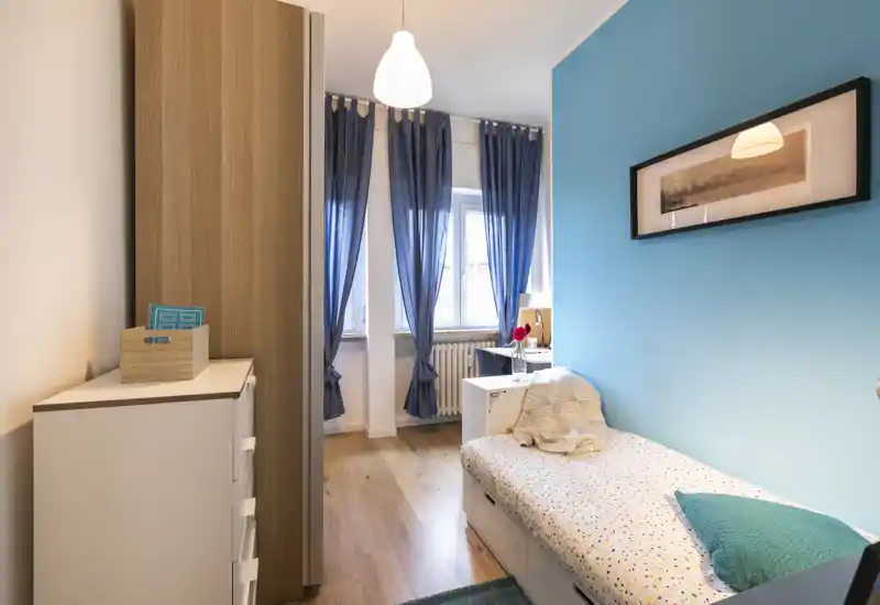 Bright, well-styled small bedroom with a single bed, bedside table and wardrobe; blue accent wall and natural light from the window create an inviting atmosphere.