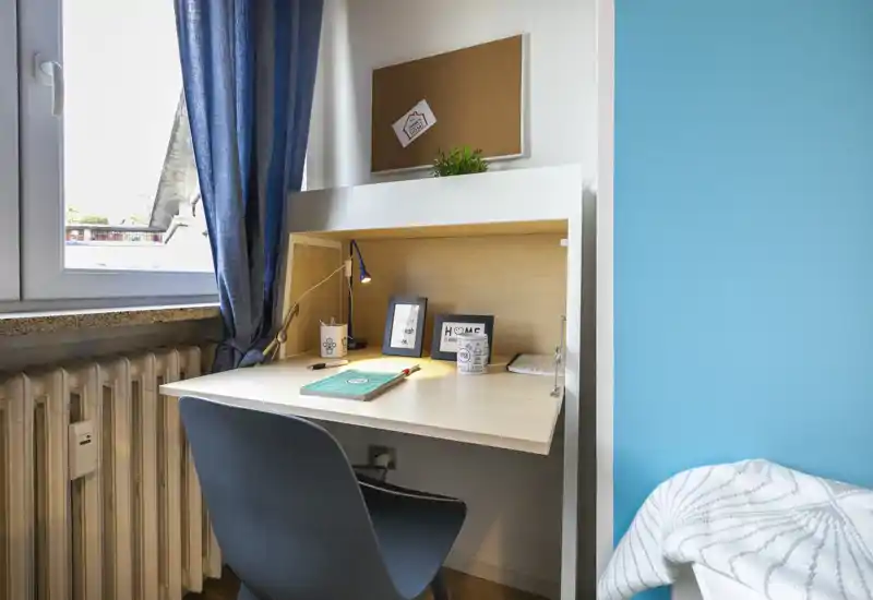 A compact bedroom corner featuring a built-in desk beside a window and a small visible portion of the bed. The workspace includes a chair, task lamp, notebooks and framed decor, in a tidy, softly lit setting.