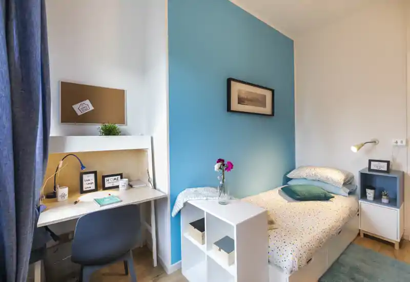 Bright, cozy single bedroom with a comfortable bed, bedside shelf and a compact desk area — ideal for work and rest. The blue accent wall and soft textiles create a welcoming, modern atmosphere.