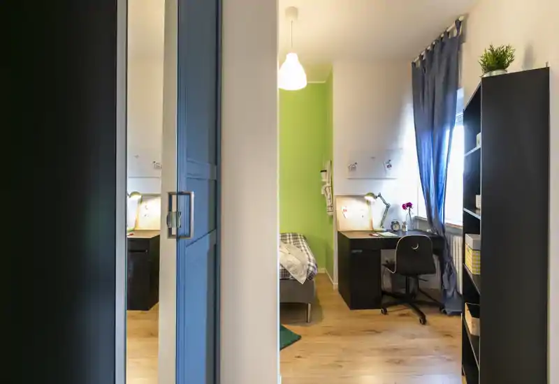 Dormitorio luminoso y ordenado con una cama individual junto a una pared verde, escritorio junto a la ventana y estanterías prácticas—ideal para estudiante o ocupante individual.