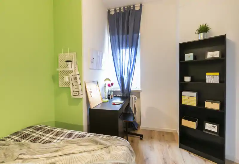Dormitorio luminoso y cuidado con cama acogedora en primer plano, escritorio junto a la ventana y estantería ordenada — ideal para estudiante o persona sola.