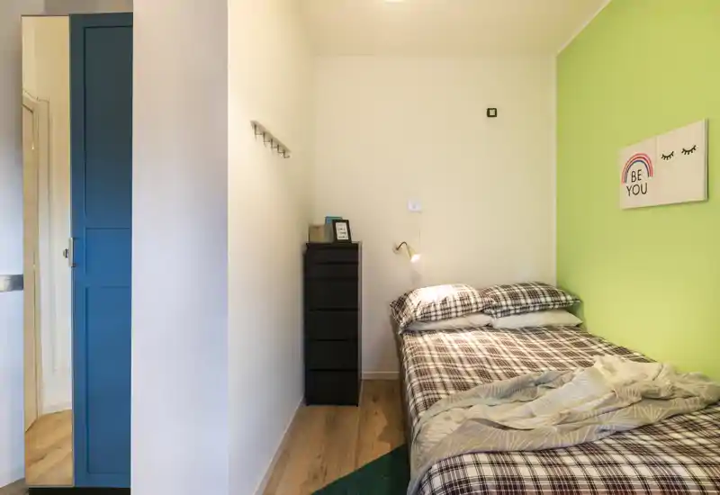 Dormitorio pequeño y luminoso con cama individual con ropa de cama estampada, cómoda estrecha, lámpara de pared y cuadro decorativo sobre una pared verde.