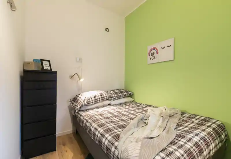 Pequeña habitación acogedora con cama doble con ropa de cama de cuadros, lámpara de lectura y una cómoda alta negra junto a una pared verde de acento.