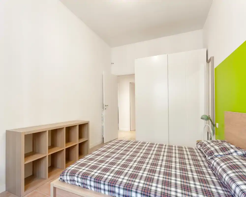 Dormitorio luminoso y minimalista con cama doble con ropa de cuadros, armario blanco y estantería sencilla. La pared verde y la lámpara de noche aportan un aire moderno y fresco.