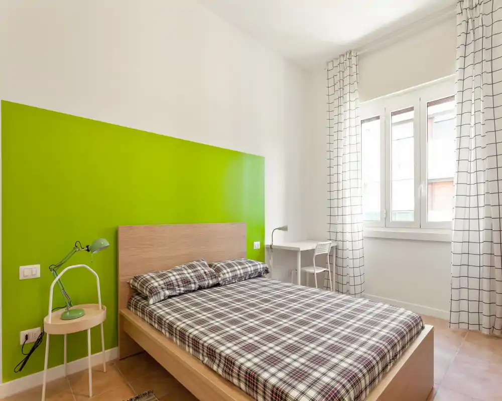 Dormitorio moderno y luminoso con cama doble, cabecero de madera y una pared verde llamativa. La luz natural entra por una gran ventana; un escritorio compacto y una lámpara de mesilla completan el conjunto funcional.