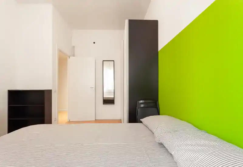 Dormitorio luminoso y minimalista con una cama grande en primer plano, una pared verde llamativa como acento y muebles negros sencillos. La imagen destaca la zona de descanso y ofrece un estilo limpio y moderno, ideal para anuncios.