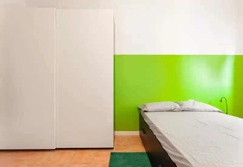 Dormitorio moderno y luminoso con cama doble frente a una pared verde brillante, amplio armario blanco y suelo de baldosa cálida — líneas limpias y decoración mínima, perfecto para el anuncio.