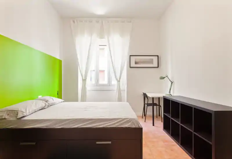Dormitorio luminoso y minimalista con cama grande, escritorio junto a la ventana y estantería baja. Paredes blancas y cortinas finas aportan una sensación fresca y aireada.