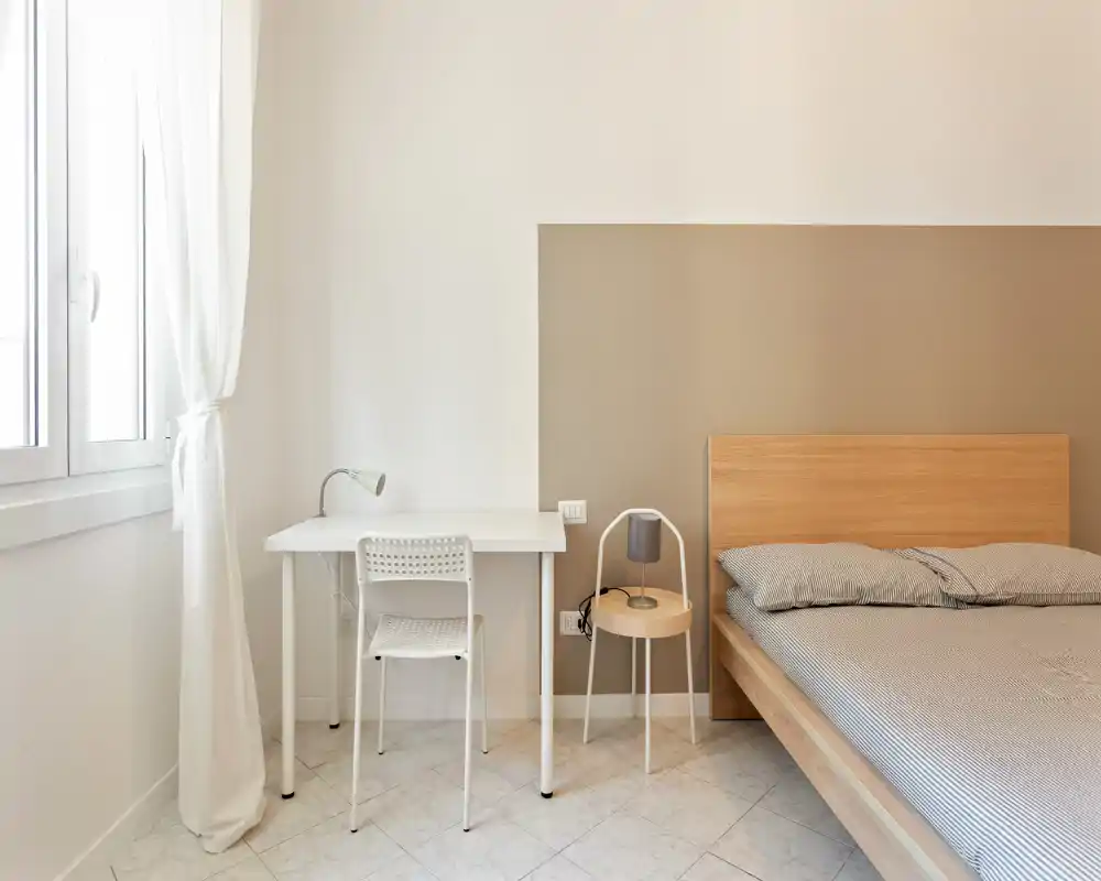 Camera da letto luminosa e minimal con struttura del letto in legno, biancheria a righe, un piccolo comodino e una scrivania compatta vicino alla finestra — perfetta per un annuncio di affitto.