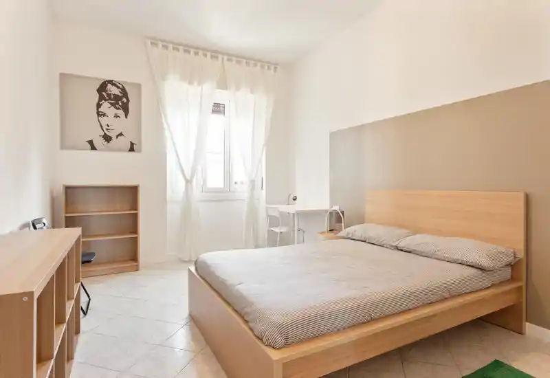 Camera da letto luminosa e minimal con letto matrimoniale in legno, biancheria neutra, una piccola scrivania vicino alla finestra e tende chiare che diffondono la luce naturale.
