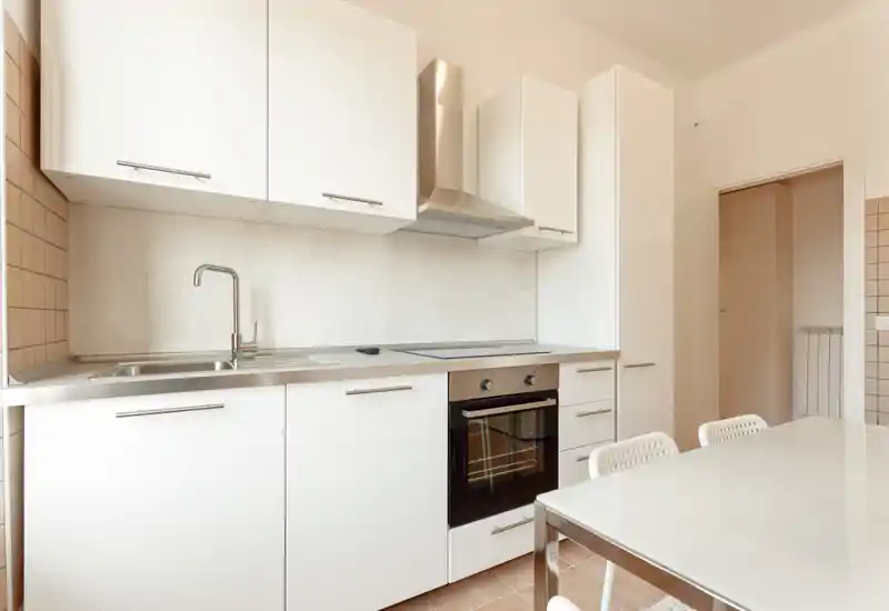 Modern white kitchen with built-in oven, sink and a dining table; bright, minimal and clean space ideal for small-group meals.