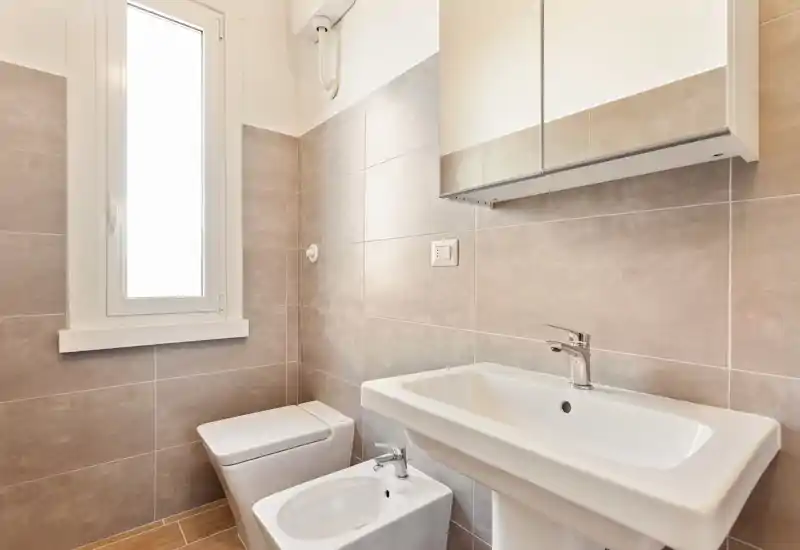 Bright, modern bathroom with a pedestal sink, bidet and toilet next to a tall frosted window; neutral tiles and a mirrored cabinet provide storage and clean lines.