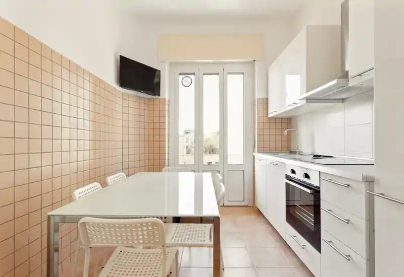 Bright modern kitchen with a glass dining table, white cabinets, built-in oven and warm tiled walls — ideal for comfortable meal prep and casual dining.