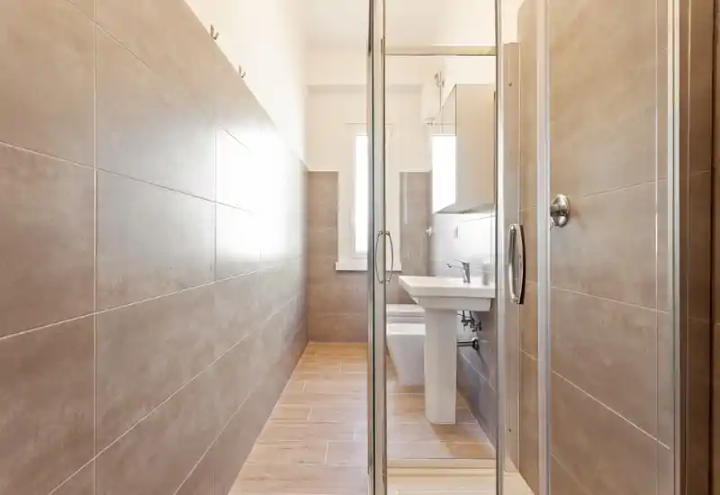 Bright, modern bathroom with a glass shower door, pedestal sink and wall-mounted toilet, finished in warm beige tiles and wood-effect flooring.