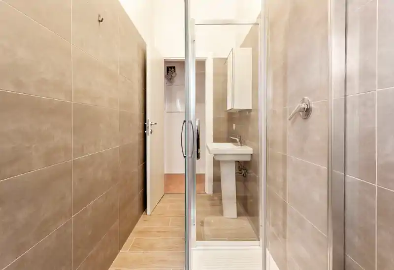 Compact modern bathroom with floor-to-ceiling neutral tiles, a pedestal sink and a glass-enclosed shower; bright, clean and minimal.