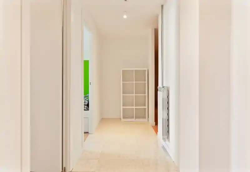 Bright, minimal hallway with tiled floor, white walls and a small shelving unit at the end — well lit and clean entrance area.