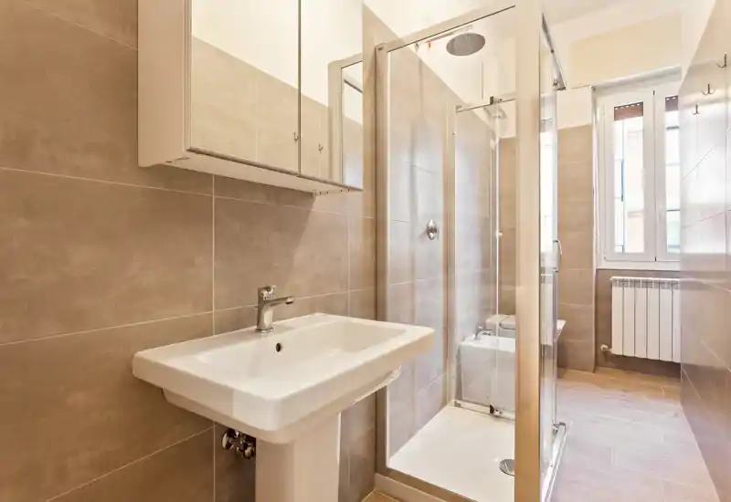 Bright, modern bathroom with a pedestal sink, glass-enclosed shower and a wall-mounted mirrored cabinet. Neutral tiles and natural light from the window create a clean, contemporary feel.