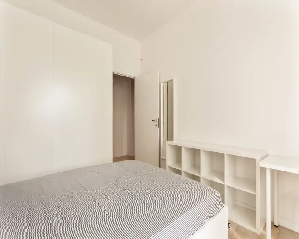 Minimal, bright bedroom with a large bed in the foreground, built-in wardrobe and simple shelving creating a clean, modern look.