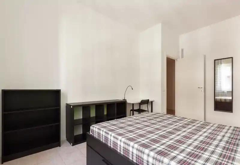 Bright, minimal bedroom with a large plaid-covered bed, simple black shelving and a small desk by the wall — clean, functional and well-lit.
