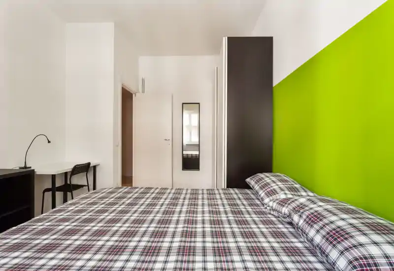 Bright, modern bedroom with a large bed dressed in patterned bedding and a vivid green accent wall; simple desk and wardrobe complete the minimalist layout.