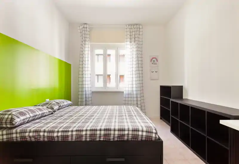 Bright, modern bedroom with a large double bed dressed in checked bedding, a green painted headboard wall and a window with patterned curtains that floods the room with natural light.