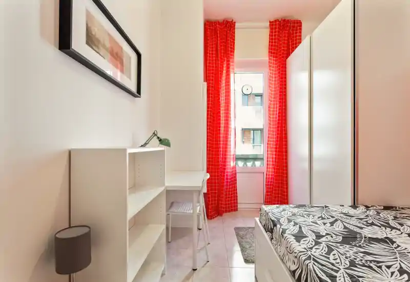 Dormitorio compacto y luminoso con cama individual estampada, estanterías blancas, un pequeño escritorio y cortinas rojas que enmarcan una ventana con luz natural.