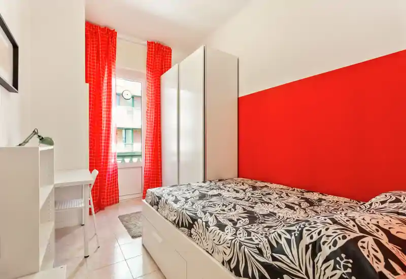 Dormitorio compacto y luminoso con una cama individual cómoda, pared roja de acento y cortinas a juego. Incluye armario blanco y pequeño escritorio, ideal para estudiante o ocupante individual.