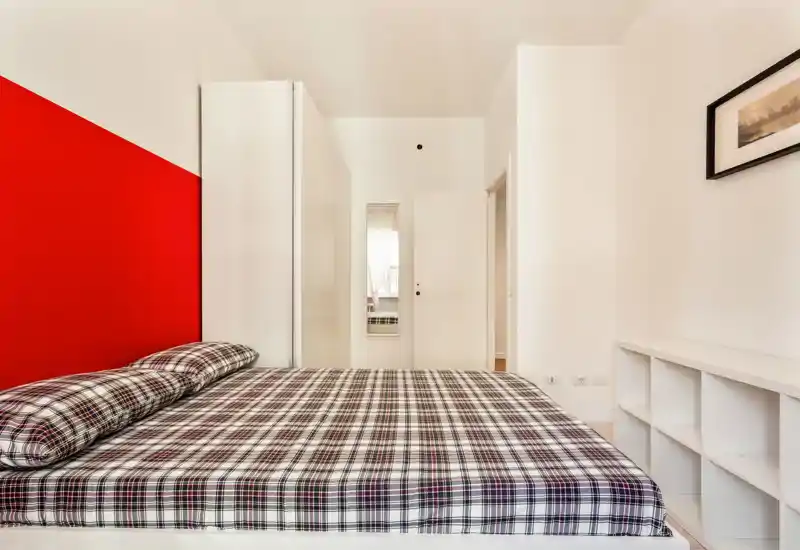 Dormitorio moderno y luminoso con una gran cama de cuadros y cabecero rojo; mobiliario minimalista y paredes blancas que crean sensación de amplitud.