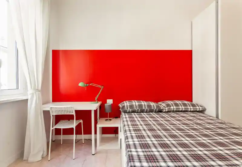 Dormitorio luminoso y compacto con cama completa, mesita de noche y pequeño escritorio junto a la ventana; pared roja y ropa de cama a cuadros que aportan un estilo moderno y acogedor.