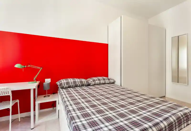 Dormitorio moderno y luminoso con una cama doble amplia con ropa de cuadros, armario blanco, pequeño escritorio y lámpara. La pared roja de acento aporta carácter y contrasta con el mobiliario minimalista blanco.