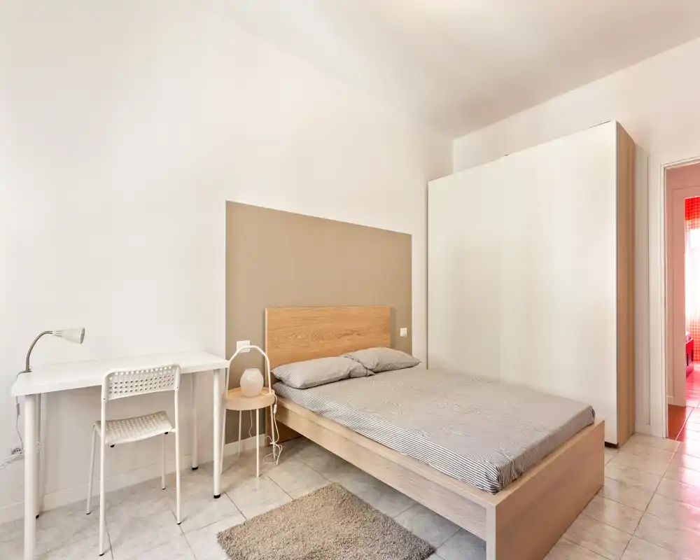 Camera da letto luminosa e moderna con letto matrimoniale, testiera in legno semplice, comodino e scrivania bianca — arredo minimal e luce naturale abbondante.