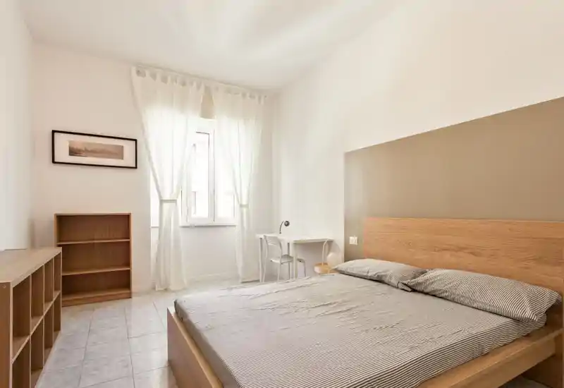Dormitorio luminoso y minimalista con cama doble, cabecero de madera y luz natural procedente de una ventana con cortinas translúcidas — estanterías sencillas y un pequeño escritorio completan la estancia.