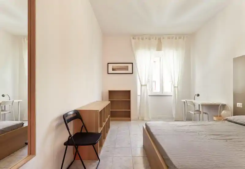 Dormitorio luminoso y minimalista con cama doble, escritorio y estanterías; la luz natural de la ventana crea un ambiente tranquilo y ordenado, ideal para estancias cortas.