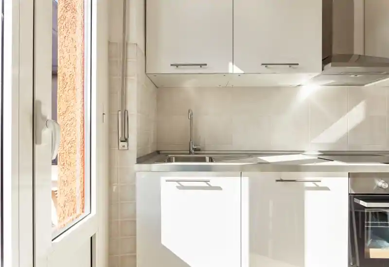 Rincón de cocina moderno y luminoso con fregadero de acero inoxidable, armarios blancos y luz natural que entra por una puerta/ventana cercana.