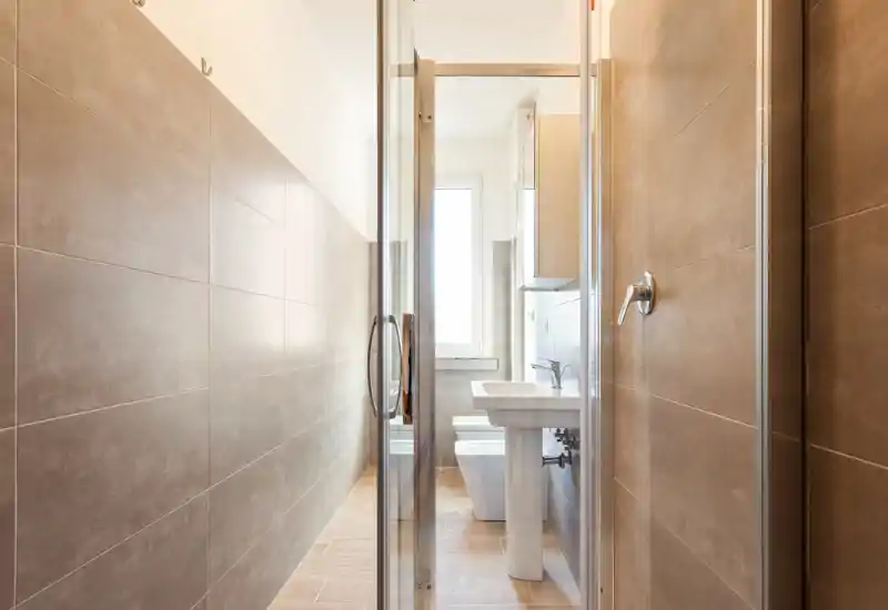 Bright, modern bathroom with a glass shower door, pedestal sink and wall-mounted fixtures. Neutral tiles and natural light create a clean, contemporary feel.
